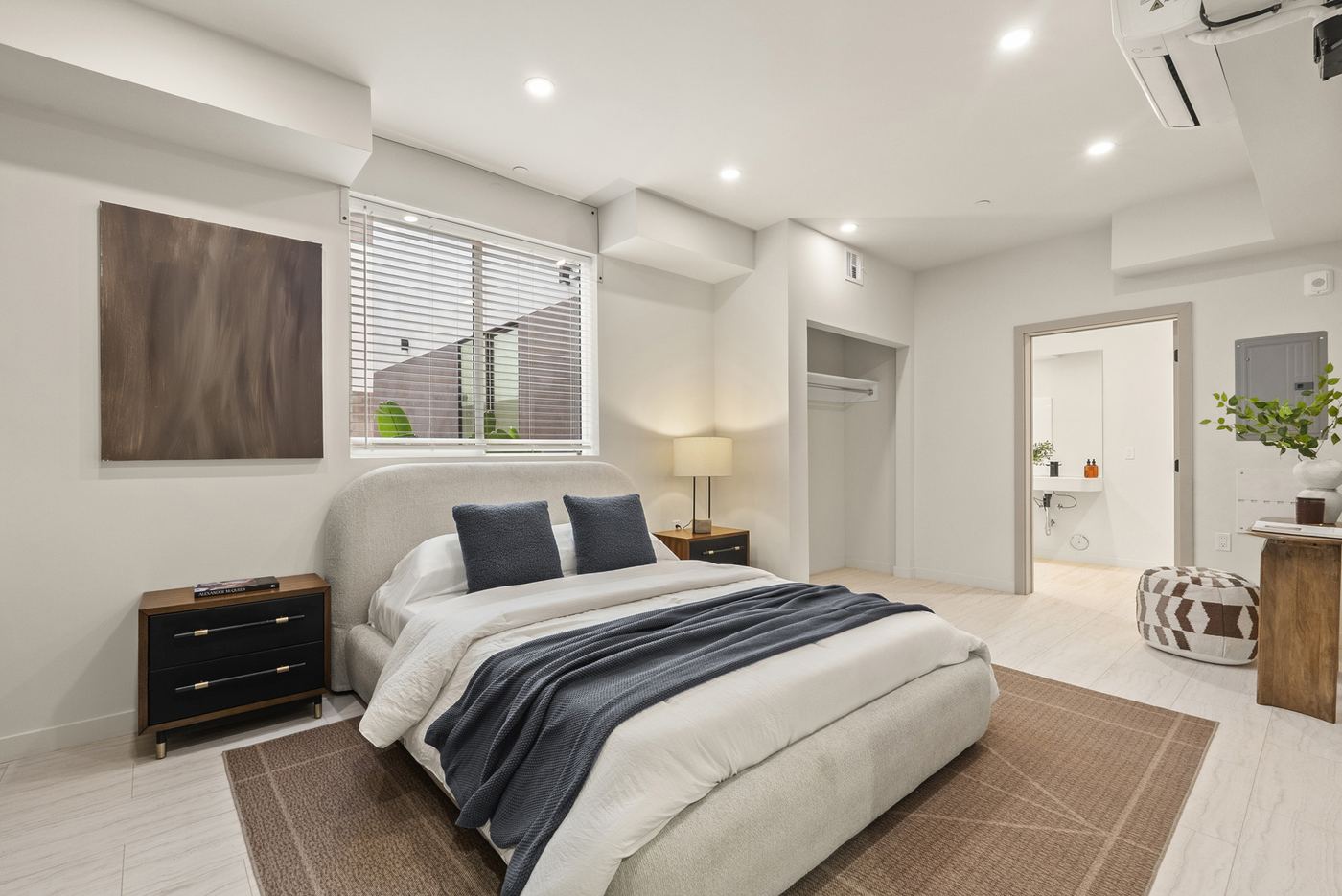 A bedroom in a ground-floor apartment. A window with blinds, a closet, and an open door to the bathroom. Furniture is staged.