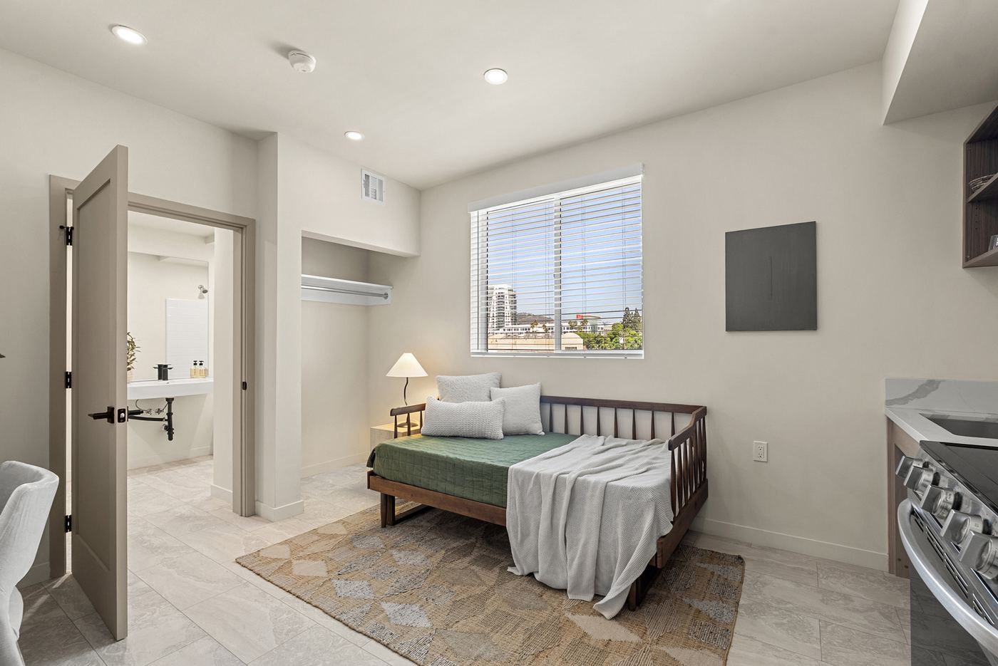 A studio apartment on an upper floor. A window, a closet, the bathroom door, and the kitchen counter at the right. Furniture is staged.