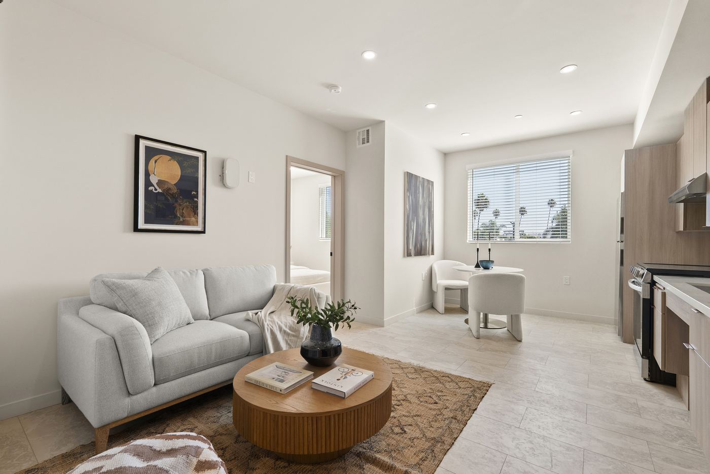 The living area of an upper-floor apartment, with a window, the kitchen on the right, and a doorway to the bedroom. Furniture is staged.